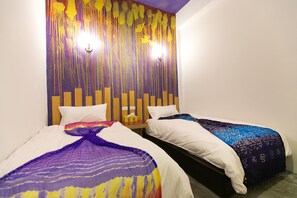 A hotel room with two beds, each with a white comforter and a colorful blanket. There is a wooden headboard with vertical slats and a wall-mounted light fixture. - Archinn (Hualien City)