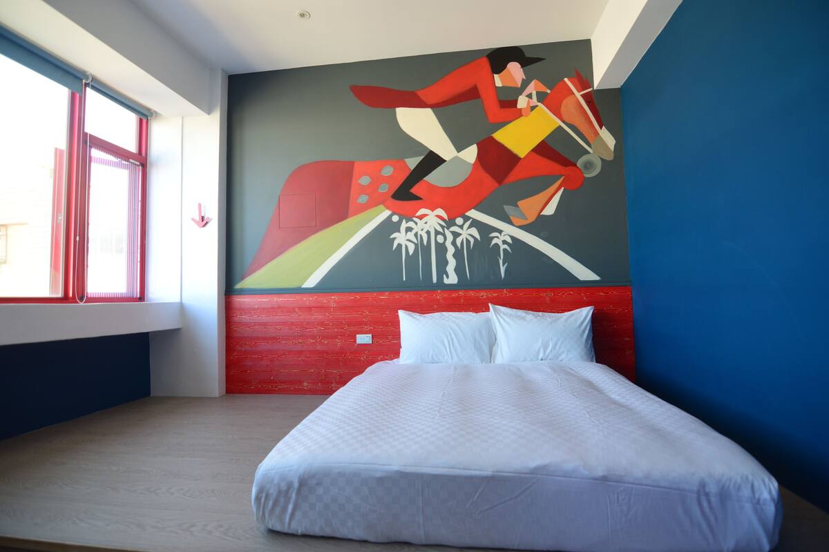 Quarto de hotel com uma cama grande, um mural na parede e uma janela com persianas vermelhas.