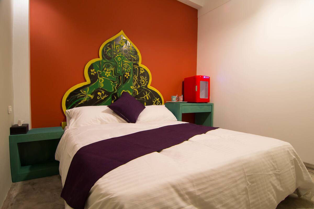 Uma cama com edredom branco e roxo, cabeceira verde com design decorativo e aquecedor vermelho sobre um criado-mudo verde.