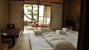 A hotel room with a bed, a desk, and a chair. There is a window with a view of trees and buildings outside. - Ryokan Kichiman (Kochi)