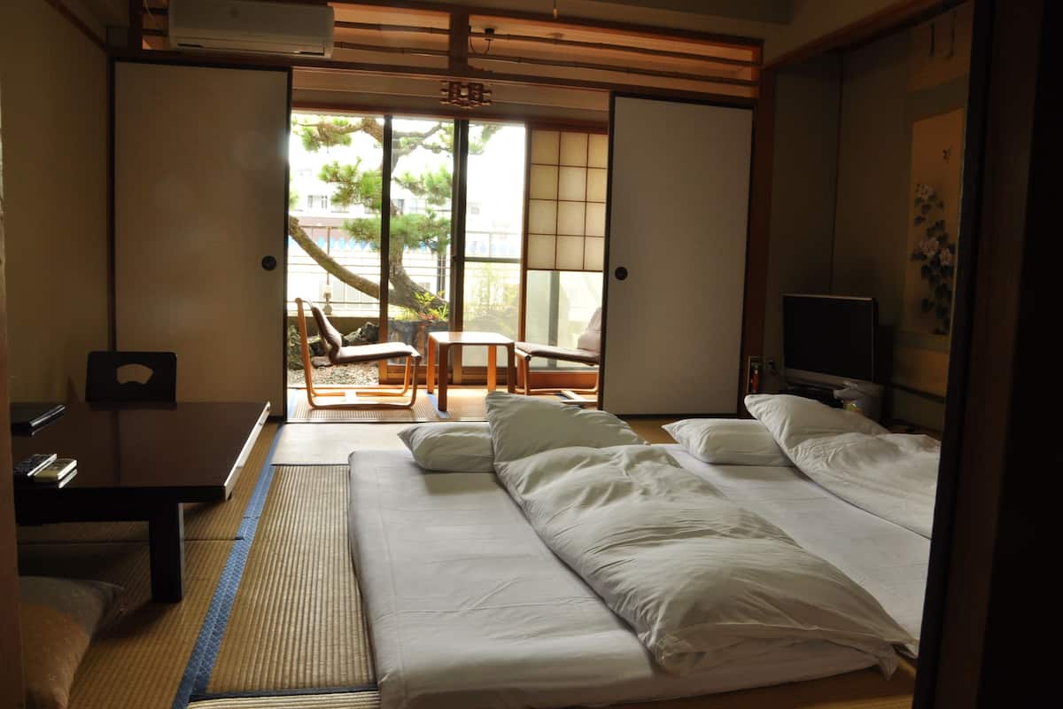 A hotel room with a bed, a desk, and a chair. There is a window with a view of trees and buildings outside.