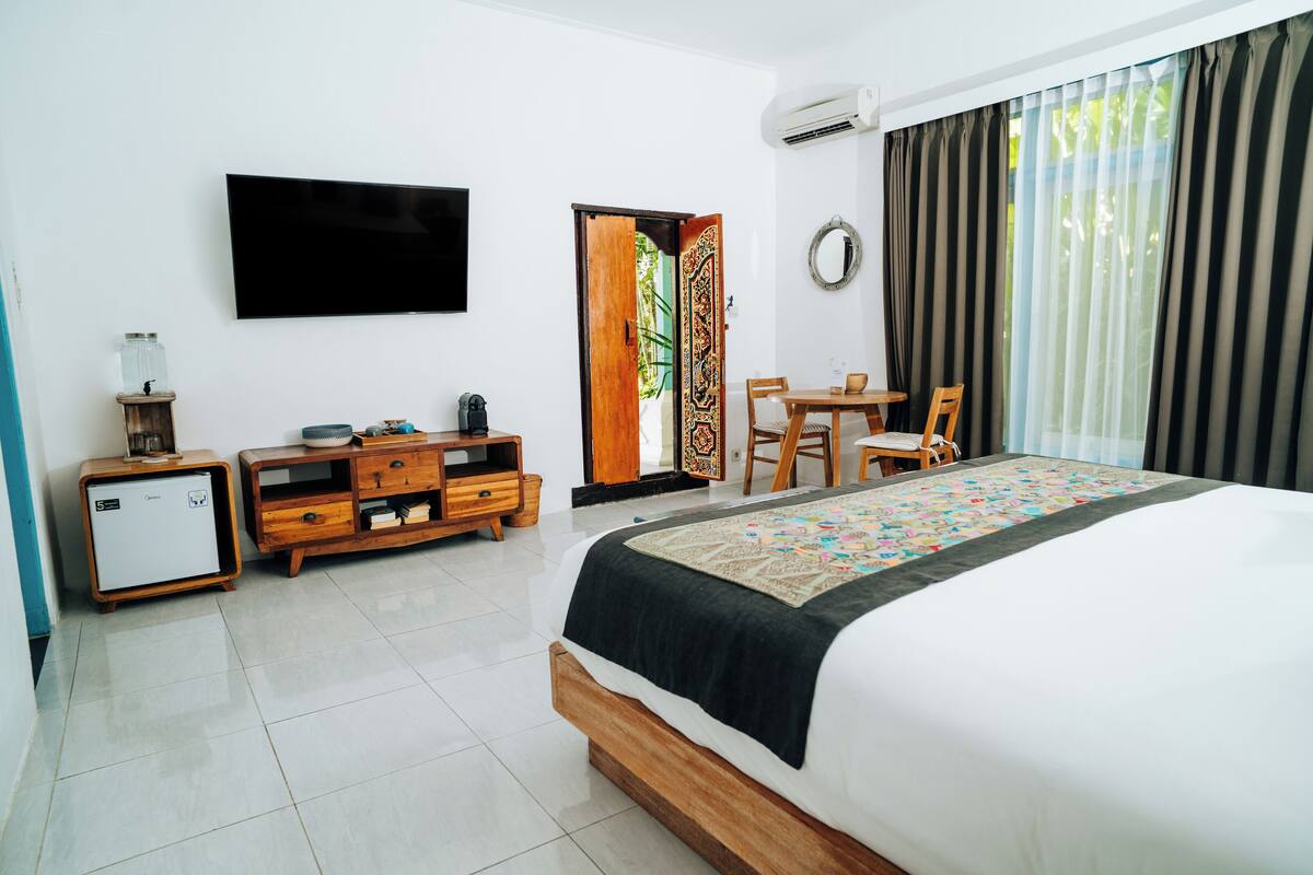 A hotel room with a bed, a desk, a chair, a TV, a small fridge, and a window with curtains.