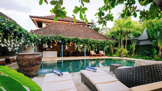 Zona junto a la piscina con sillones de descanso, una pérgola con luces colgantes y una casa al fondo.