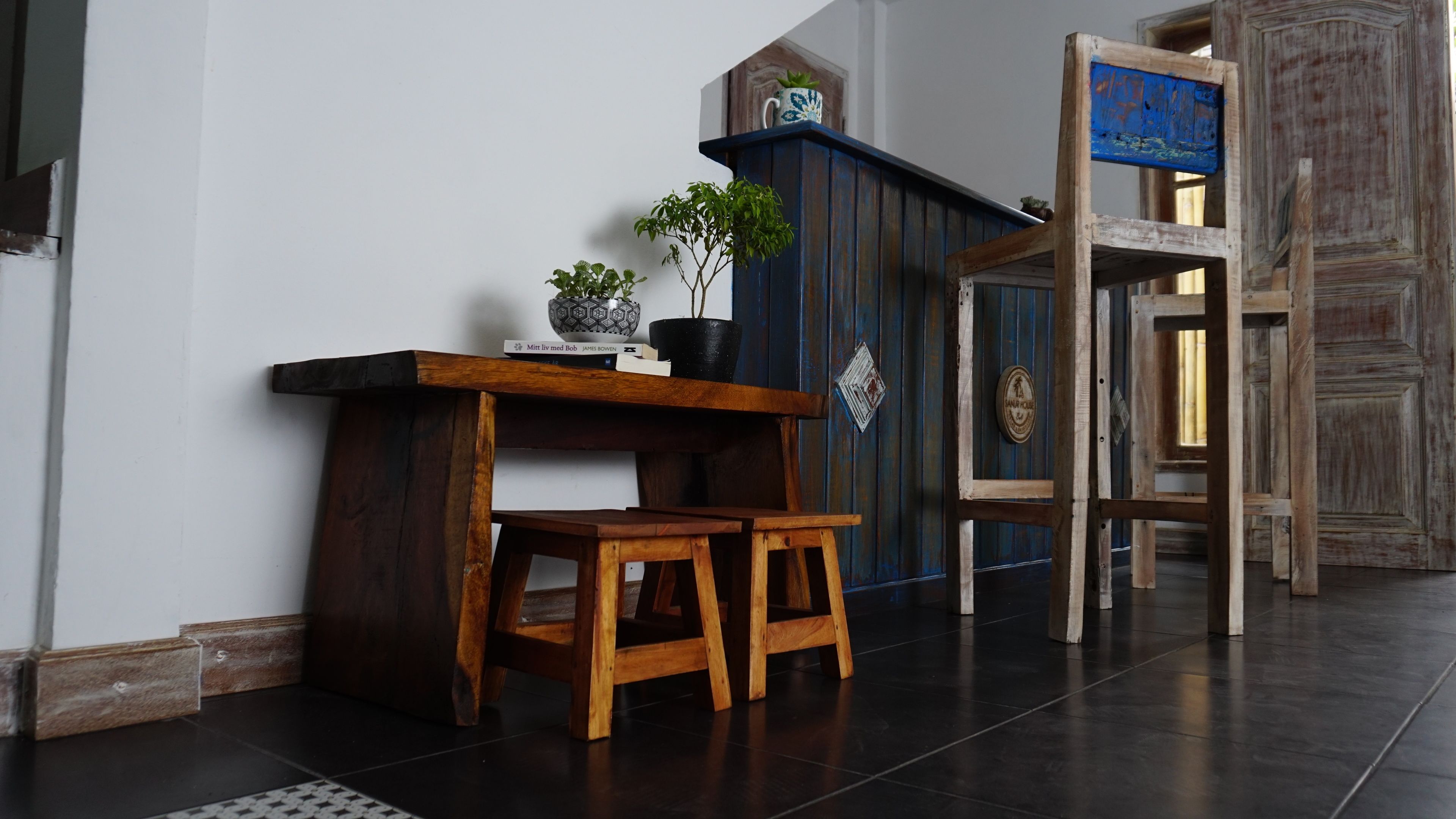 A wooden bench with a small table and two stools in a hallway with dark flooring and a blue door.