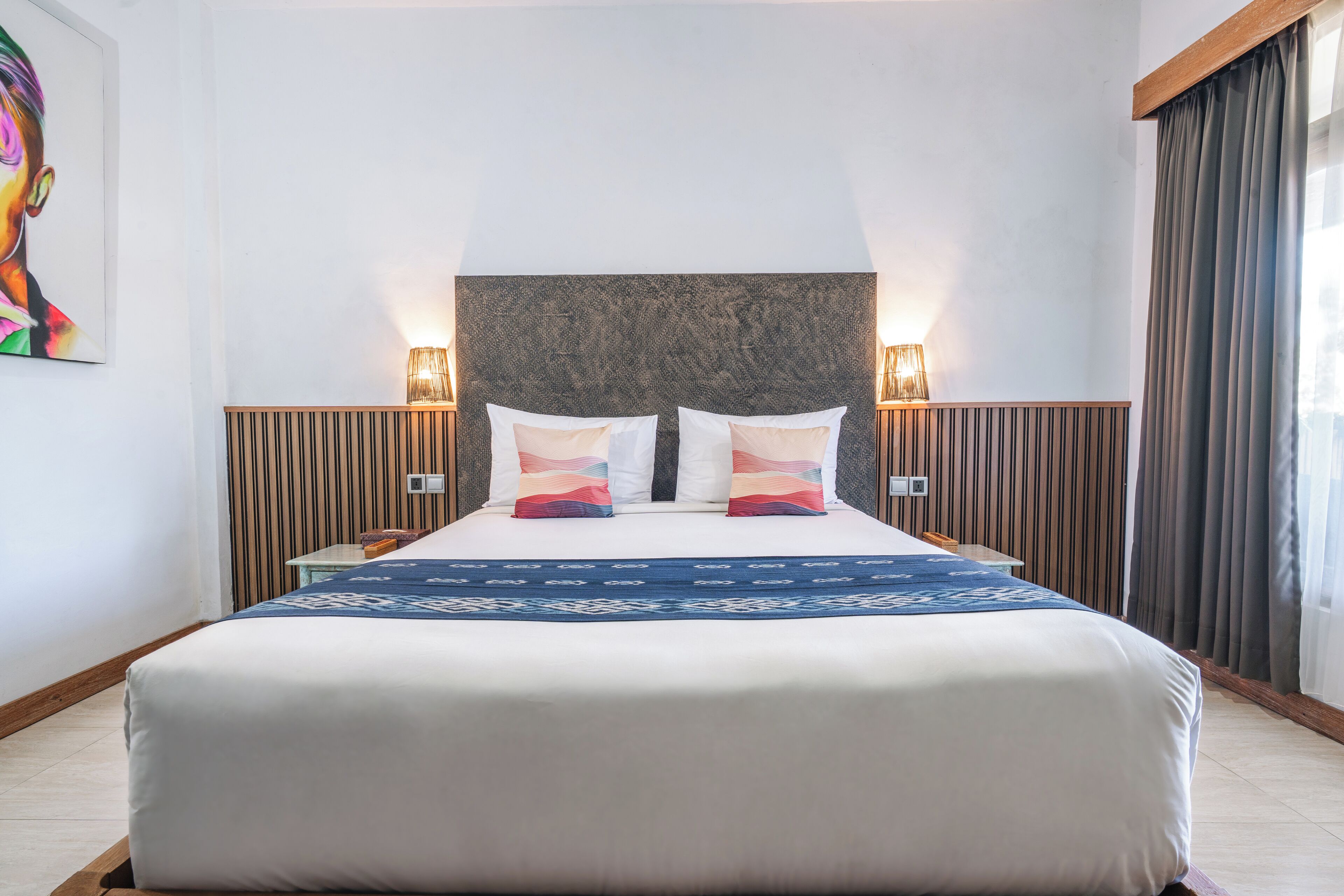 A hotel room with a large bed, two bedside lamps, a wooden headboard, and a colorful painting on the wall.