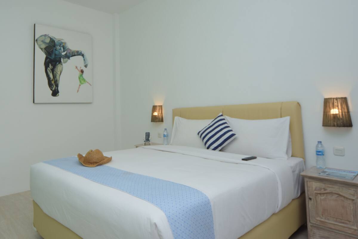 A neatly made bed with a blue and white striped pillow, a brown hat on the bed, and a painting of an elephant and a child on the wall.