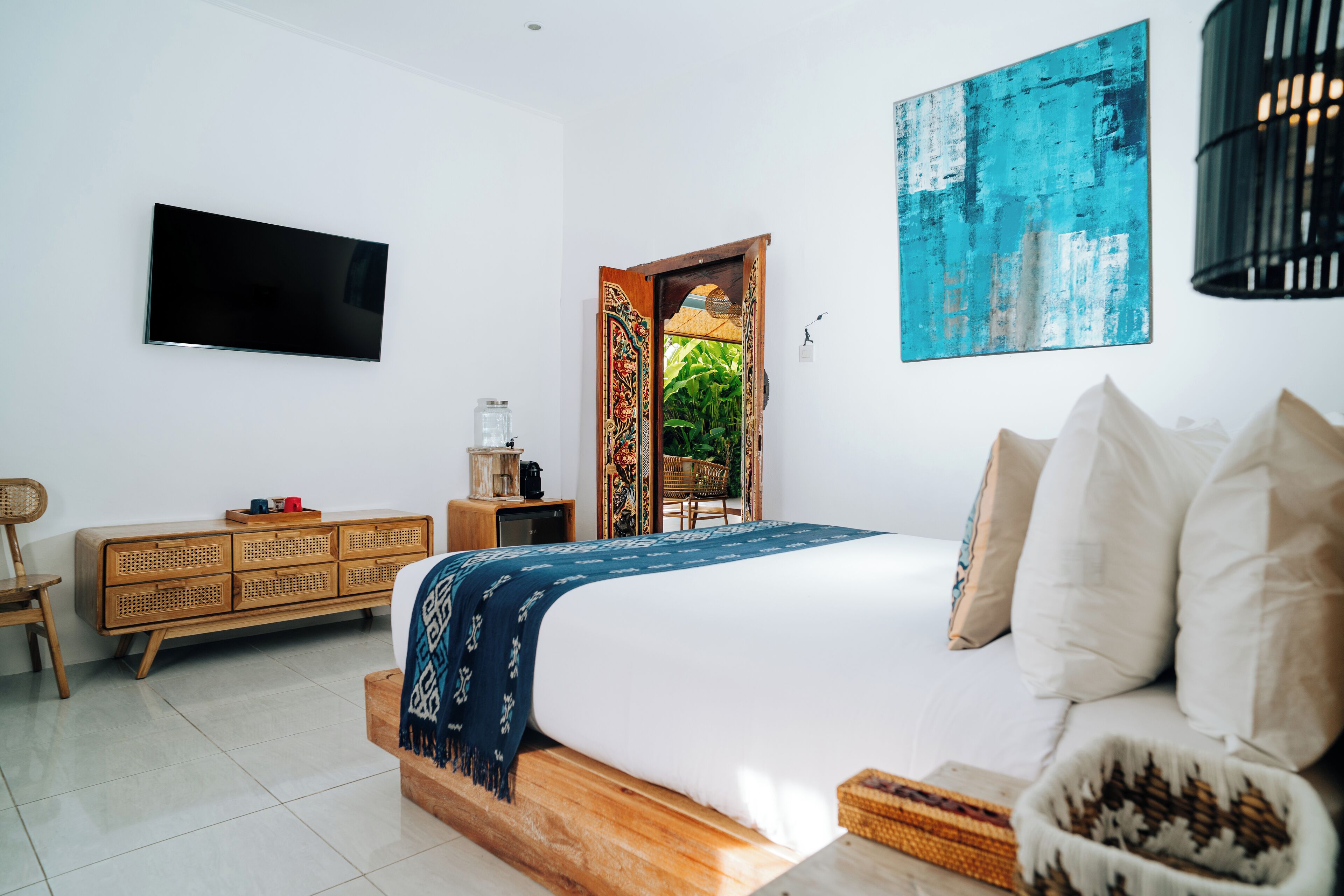 A modern bedroom with a large bed, a television, a wooden dresser, and a view of the outdoors through an open door.