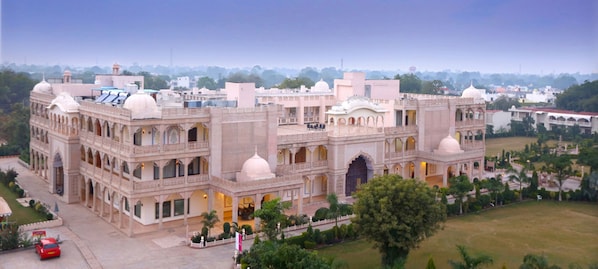 A large, historic building with multiple domes and arches, surrounded by greenery and other buildings in the background.