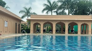Zona de piscina con una persona sosteniendo una bebida, palmeras y un edificio con arcos al fondo.