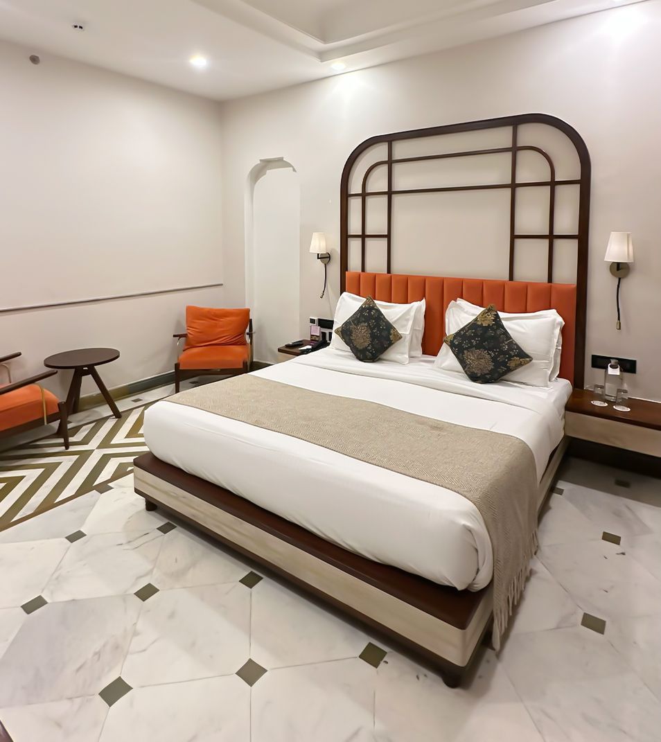 A hotel room with a large bed, a small table, an orange chair, and a patterned floor.