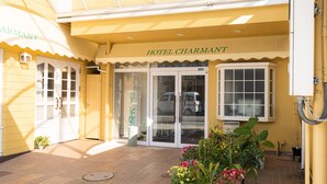 The entrance of Hotel Charmant with a tiled pavement, potted plants, and a vehicle parked outside.