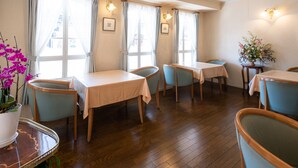A hotel dining area with wooden tables and chairs, large windows with curtains, and a potted plant.