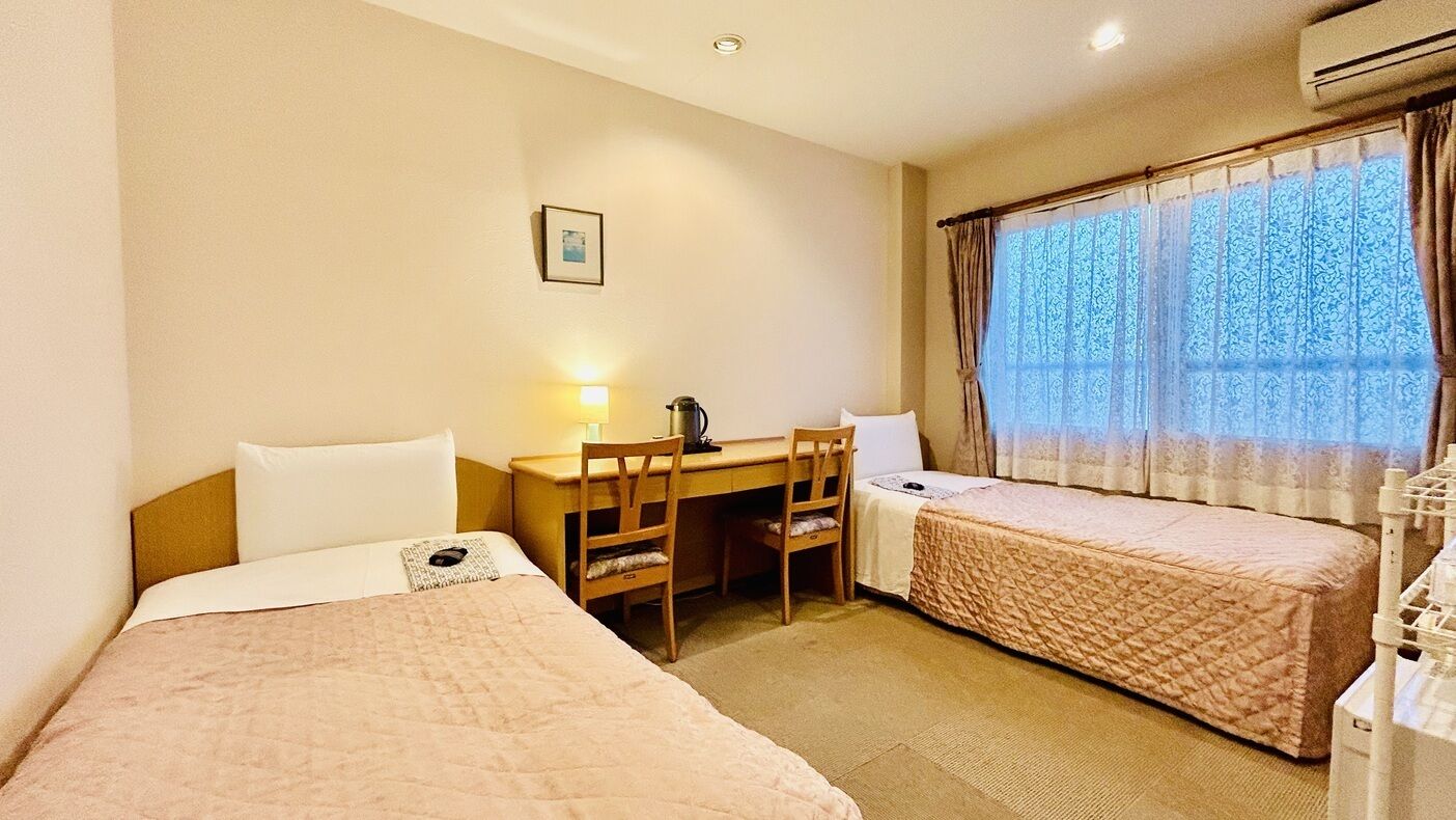 Ein Hotelzimmer mit zwei Betten, einem Schreibtisch, einem Stuhl, einer Lampe und einem Vorhangfenster.