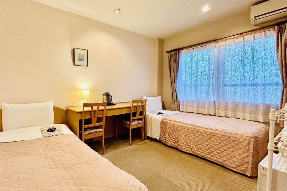 Une chambre d’hôtel avec deux lits, un bureau, une chaise, une lampe et une fenêtre avec des rideaux.