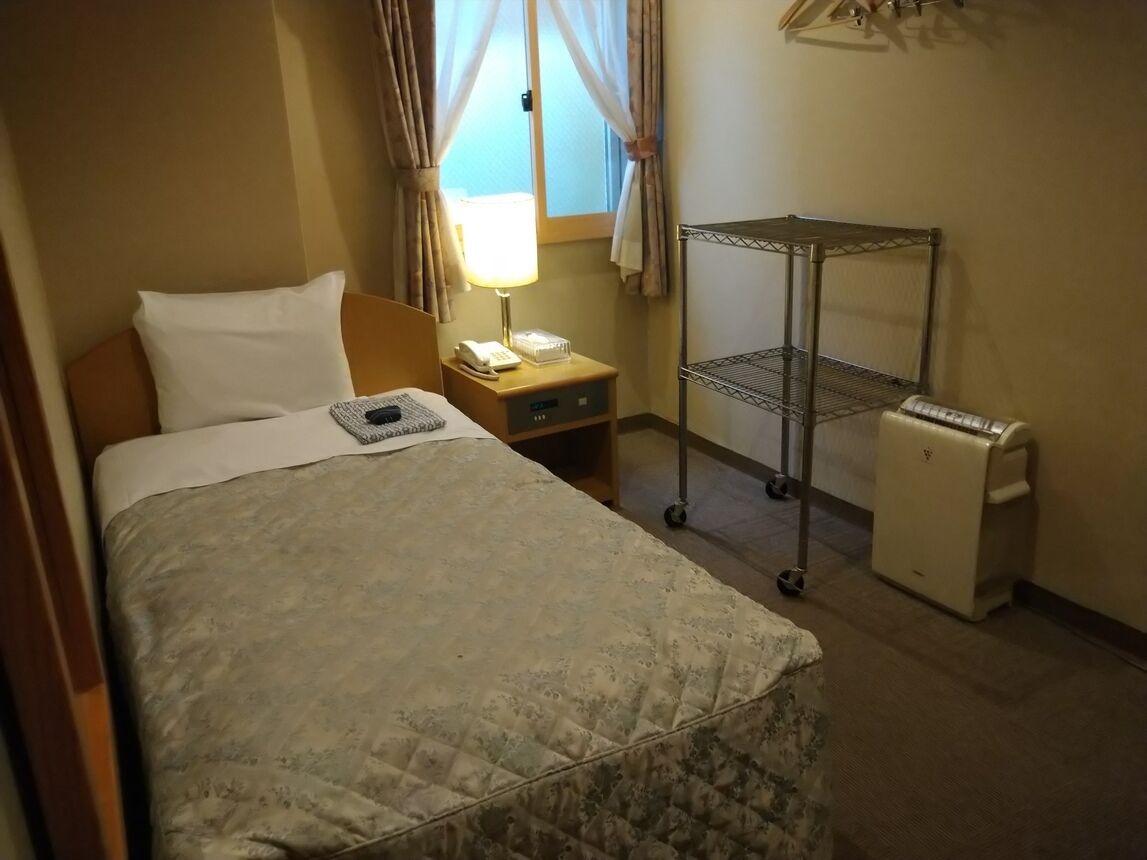 Ein Hotelzimmer mit einem Bett, einem Nachttisch mit Lampe, einem kleinen Schreibtisch, einem Metallregal und einem kompakten Kühlschrank.