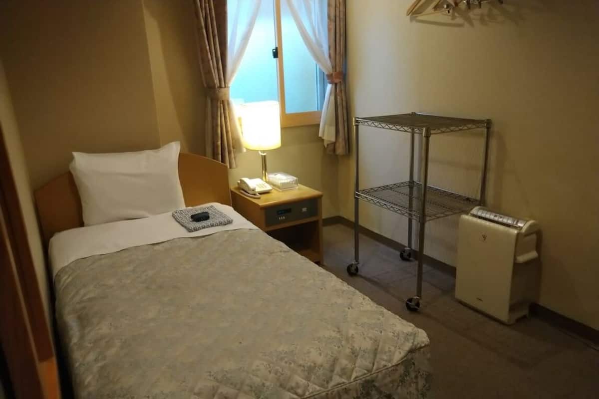 Une chambre d’hôtel comprenant un lit, une table de chevet avec une lampe, un petit bureau, une étagère en métal et un mini-réfrigérateur.