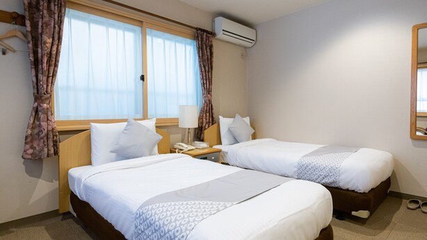 A hotel room with two beds, a bedside table with a lamp, an air conditioning unit, and a window with curtains.