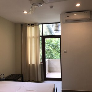 A hotel room with a bed, a window with curtains, an air conditioning unit, and a view of greenery outside.