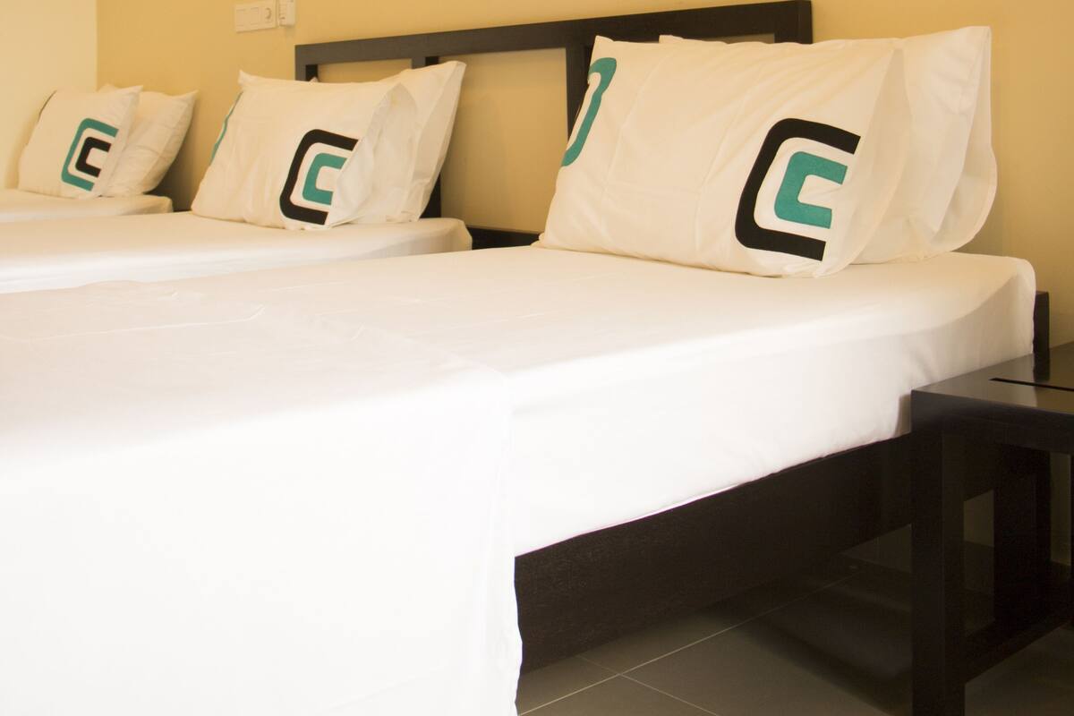 A bed with white bedding and pillows featuring a green and black logo, a dark wooden headboard, and a small bedside table.