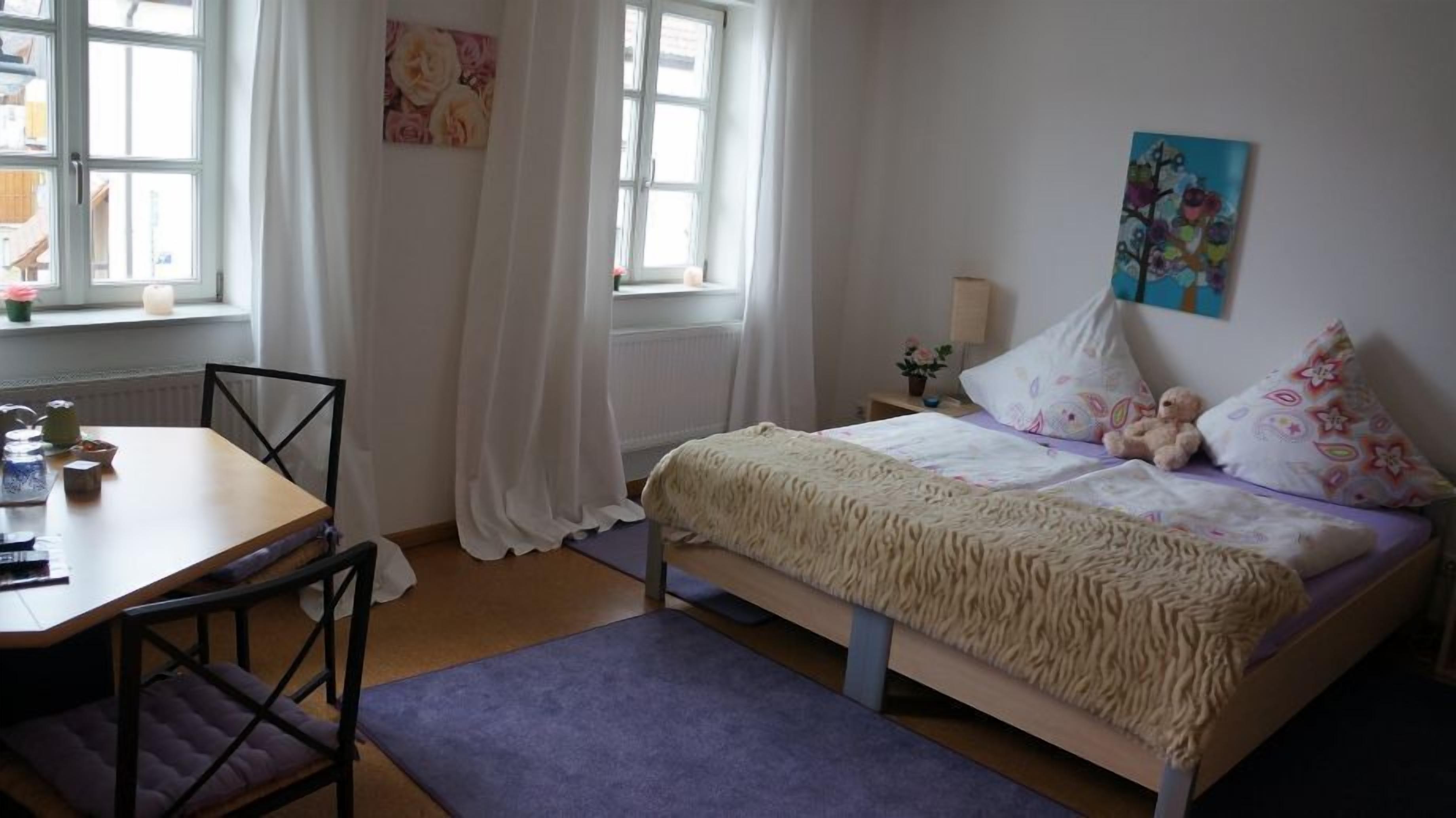 Un dormitorio con cama, escritorio y silla. Se ve una ventana con cortinas, un radiador y una pared con un cuadro.
