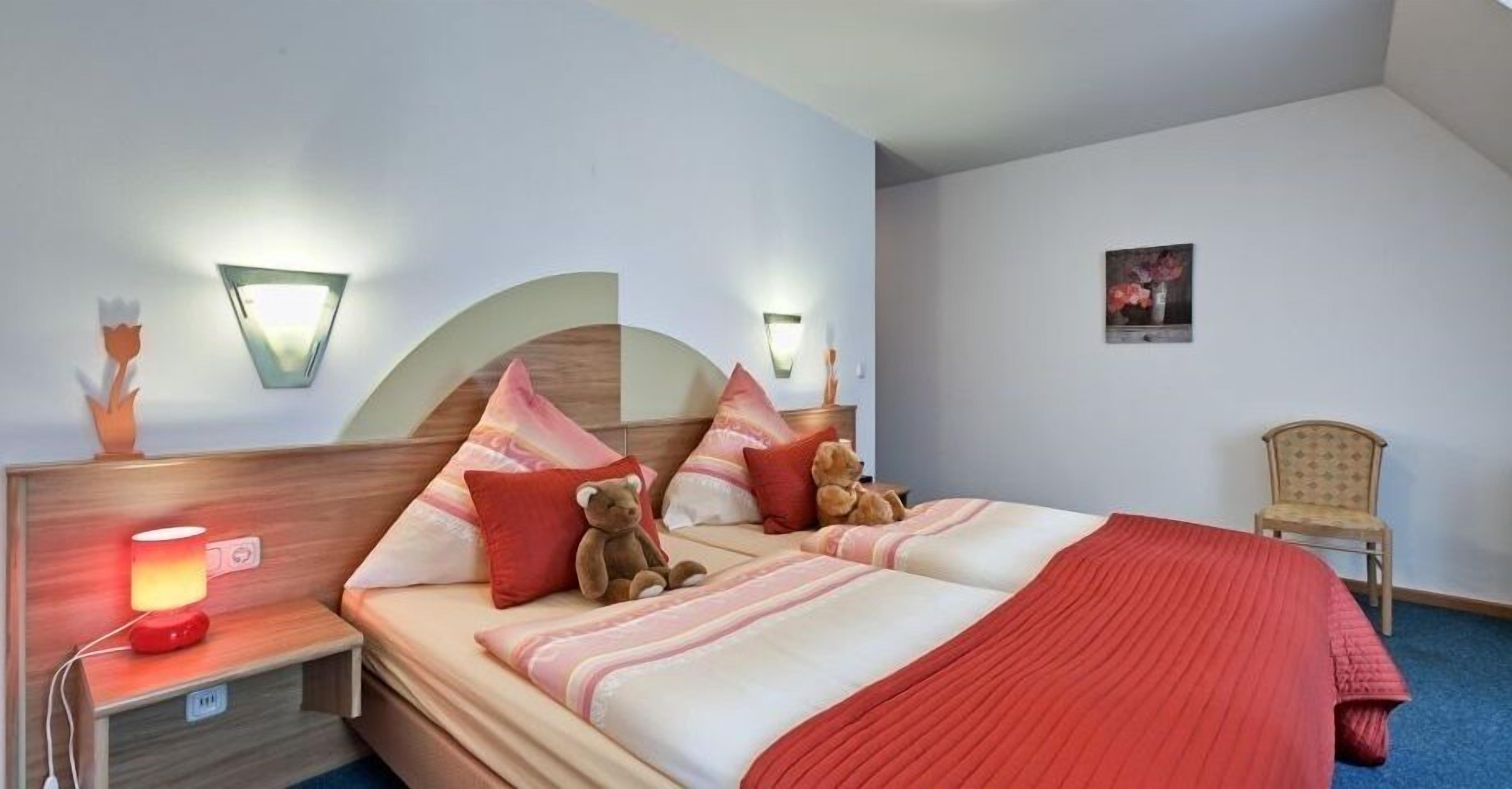 Quarto com cama, almofadas, um urso de pelúcia, mesa de cabeceira com abajur e uma cadeira.