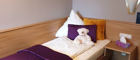 Un lit bien fait, avec une couverture violette, un oreiller blanc et un ours en peluche. Une petite table de chevet est placée à côté du lit, sur laquelle se trouve une boîte de mouchoirs. Un miroir est accroché au mur.