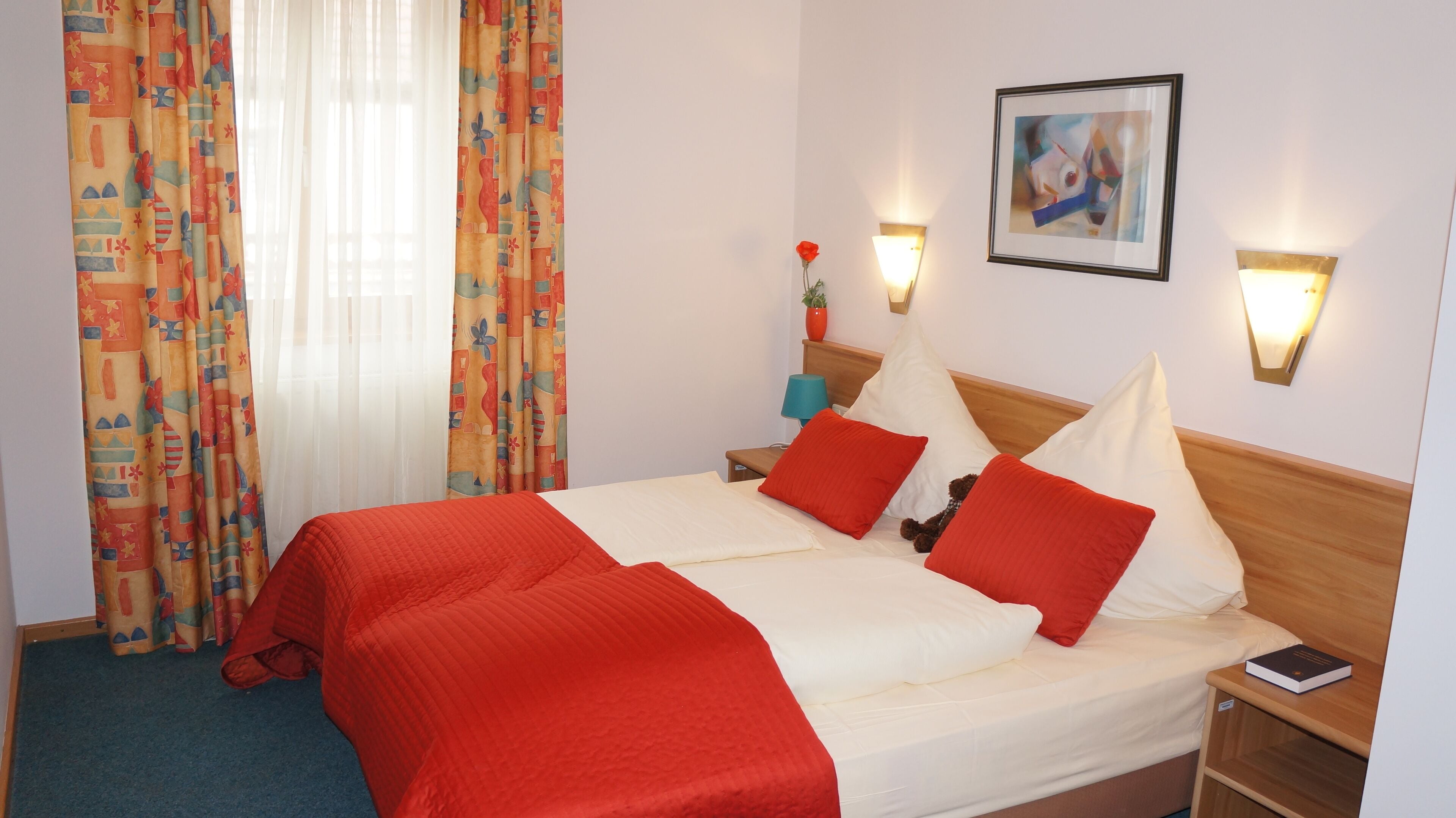 A hotel room with a bed, two bedside tables, a nightstand, a lamp, a framed picture on the wall, and a window with curtains.