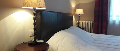 A hotel room with a bed, two bedside lamps, a nightstand, a radiator, and a window with curtains.