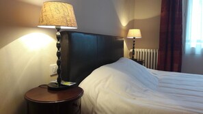 A hotel room with a bed, two bedside lamps, a nightstand, a radiator, and a window with curtains. - Hôtel les Aubépines (Illiers-Combray)