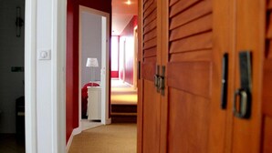 Un pasillo con una puerta de madera, una pared blanca y una pared roja. Se ve una cama con una colcha roja y una lámpara blanca al fondo.