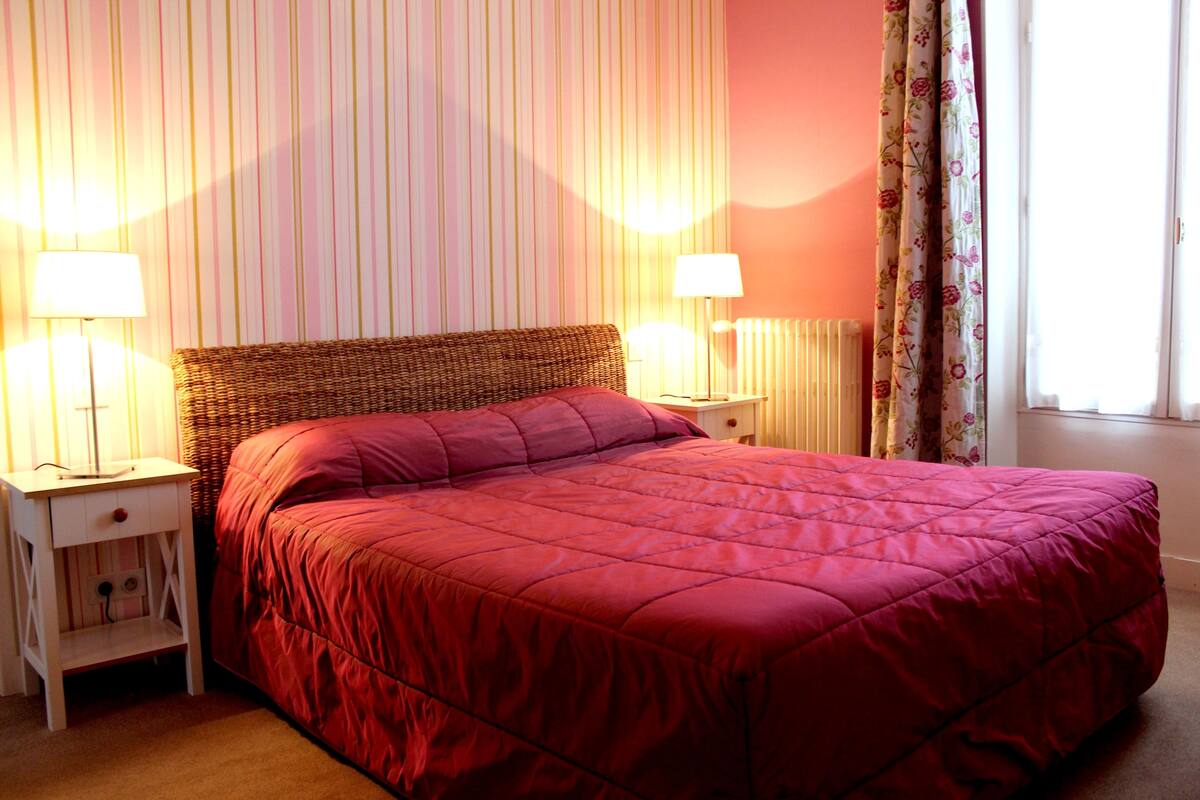 A neatly made bed with a red quilt in a room with striped wallpaper and two bedside lamps.