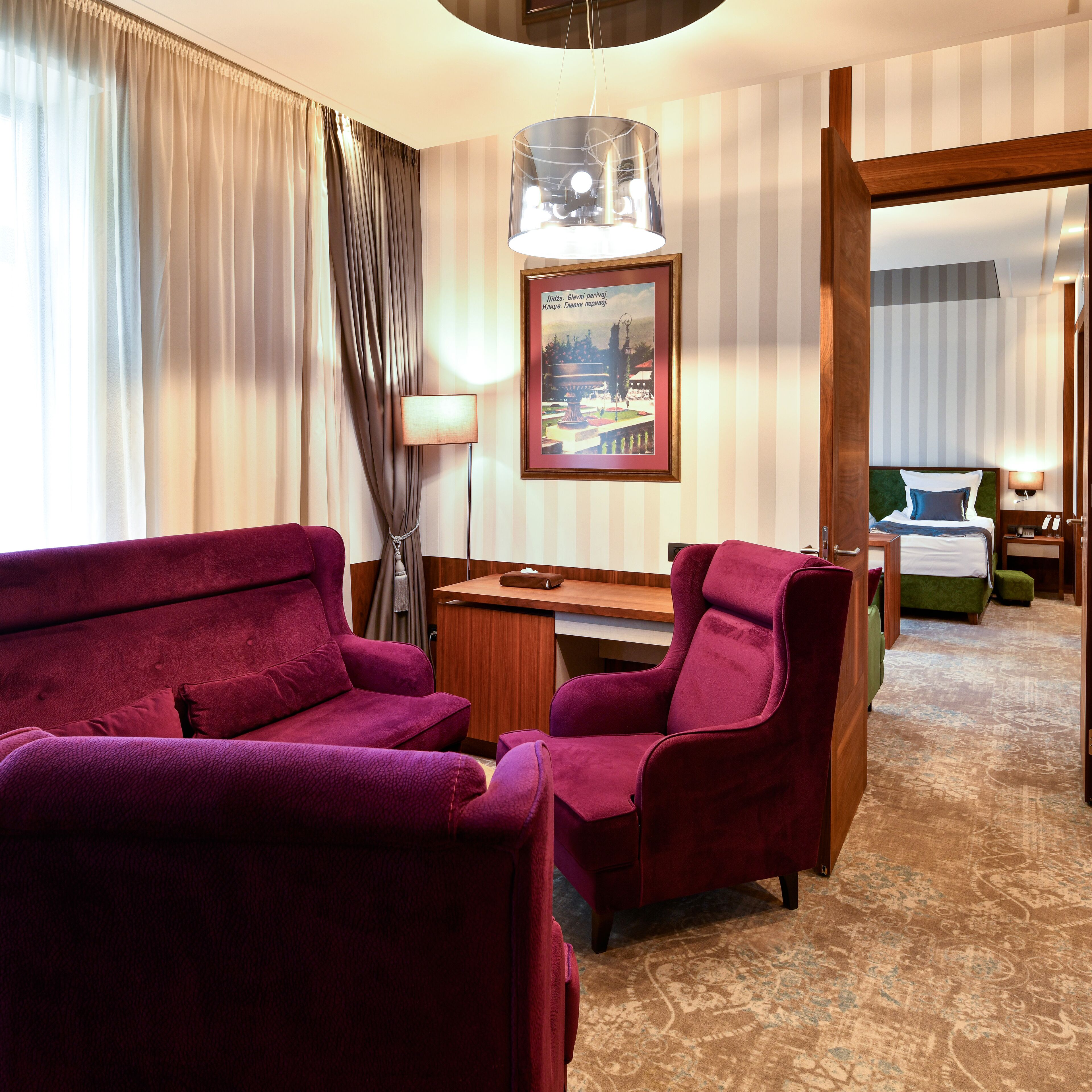 Une chambre d’hôtel avec une grande fenêtre, un bureau, deux fauteuils violets, un lit et un tableau encadré au mur.