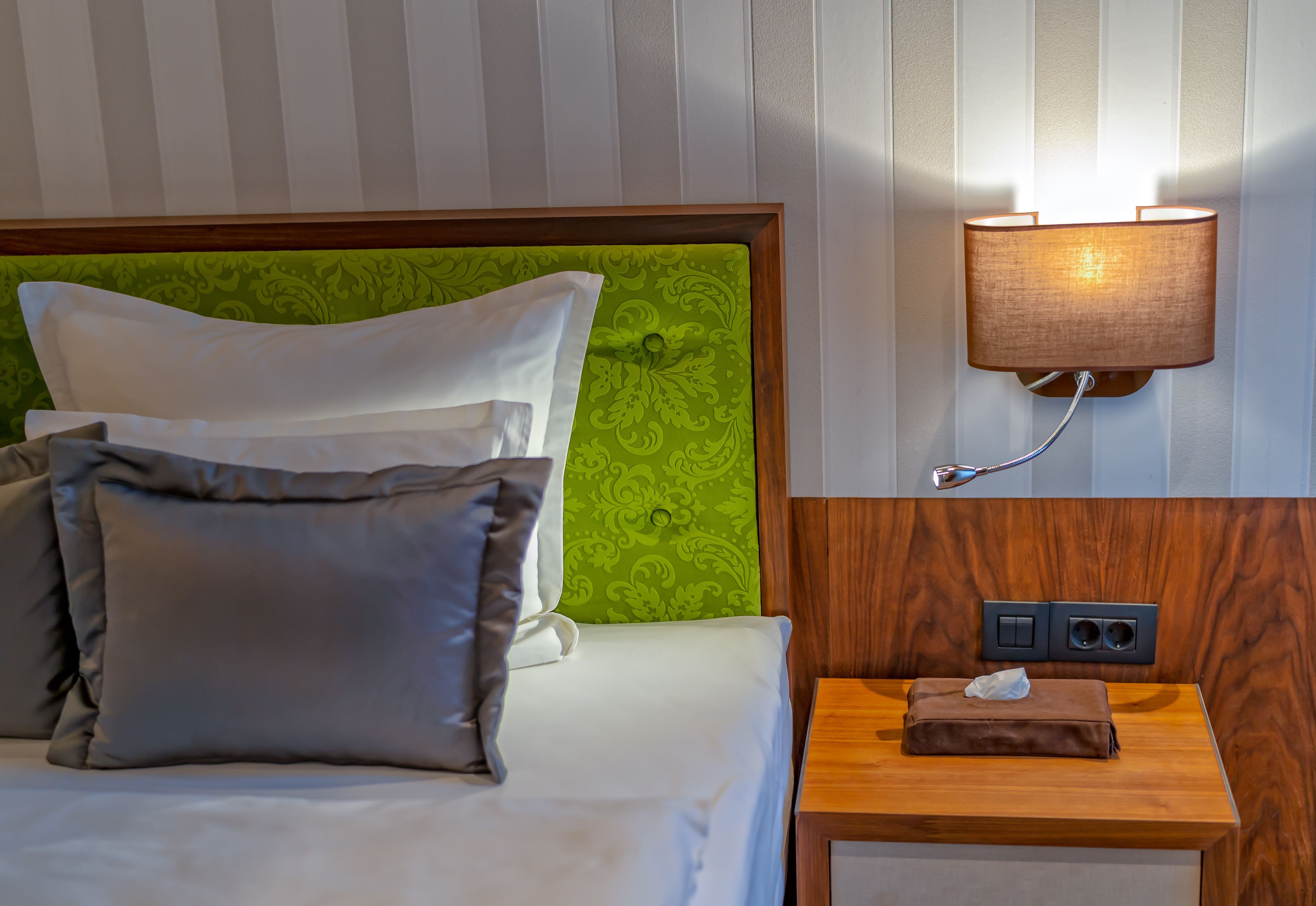 Un lit bien fait, avec une tête de lit verte, une lampe de chevet et une table de chevet en bois sur laquelle se trouve une boîte de mouchoirs.