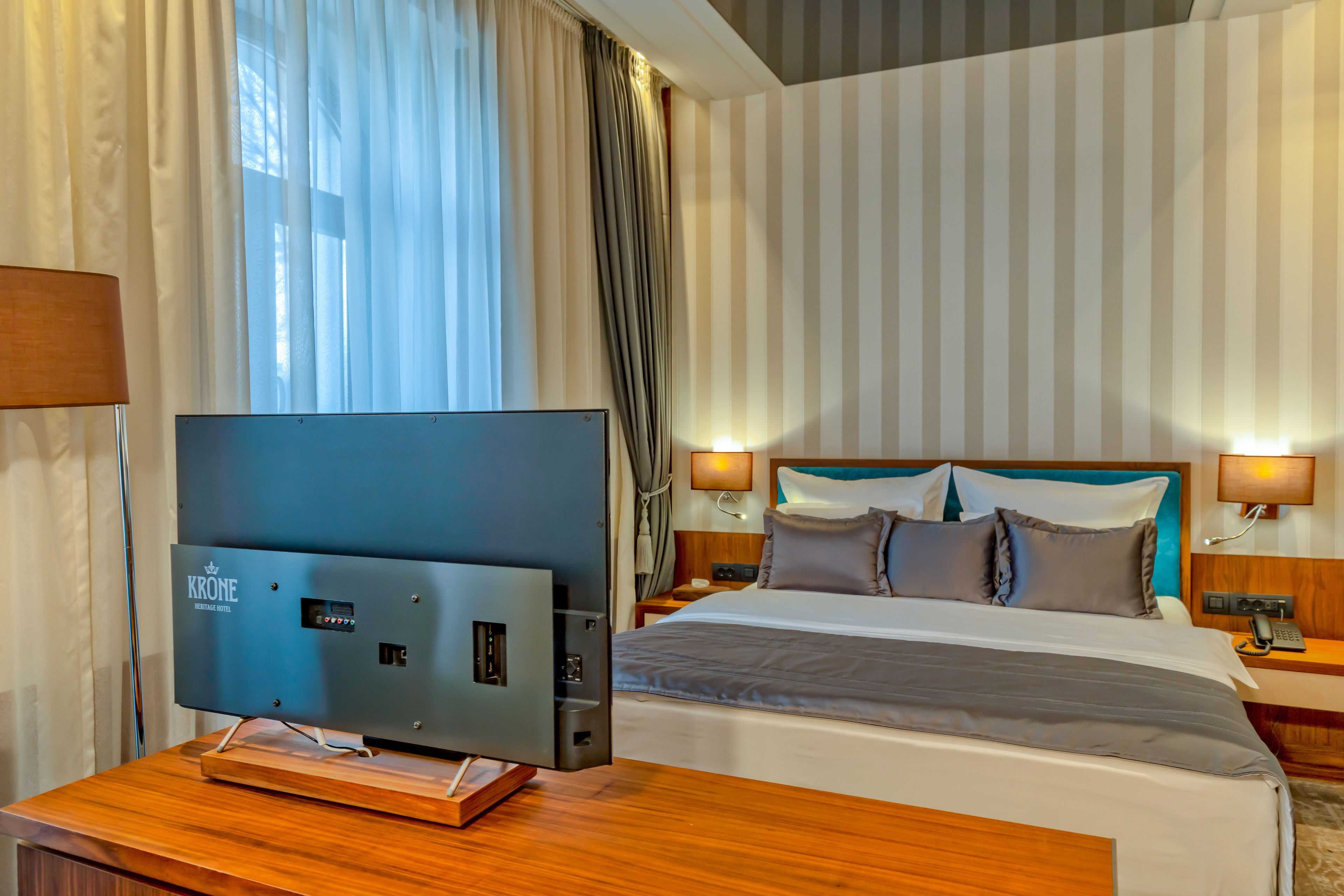 Habitación de hotel con un televisor de pantalla plana grande sobre una mesa de madera, una cama con muchas almohadas, dos lámparas de noche y una pared a rayas.