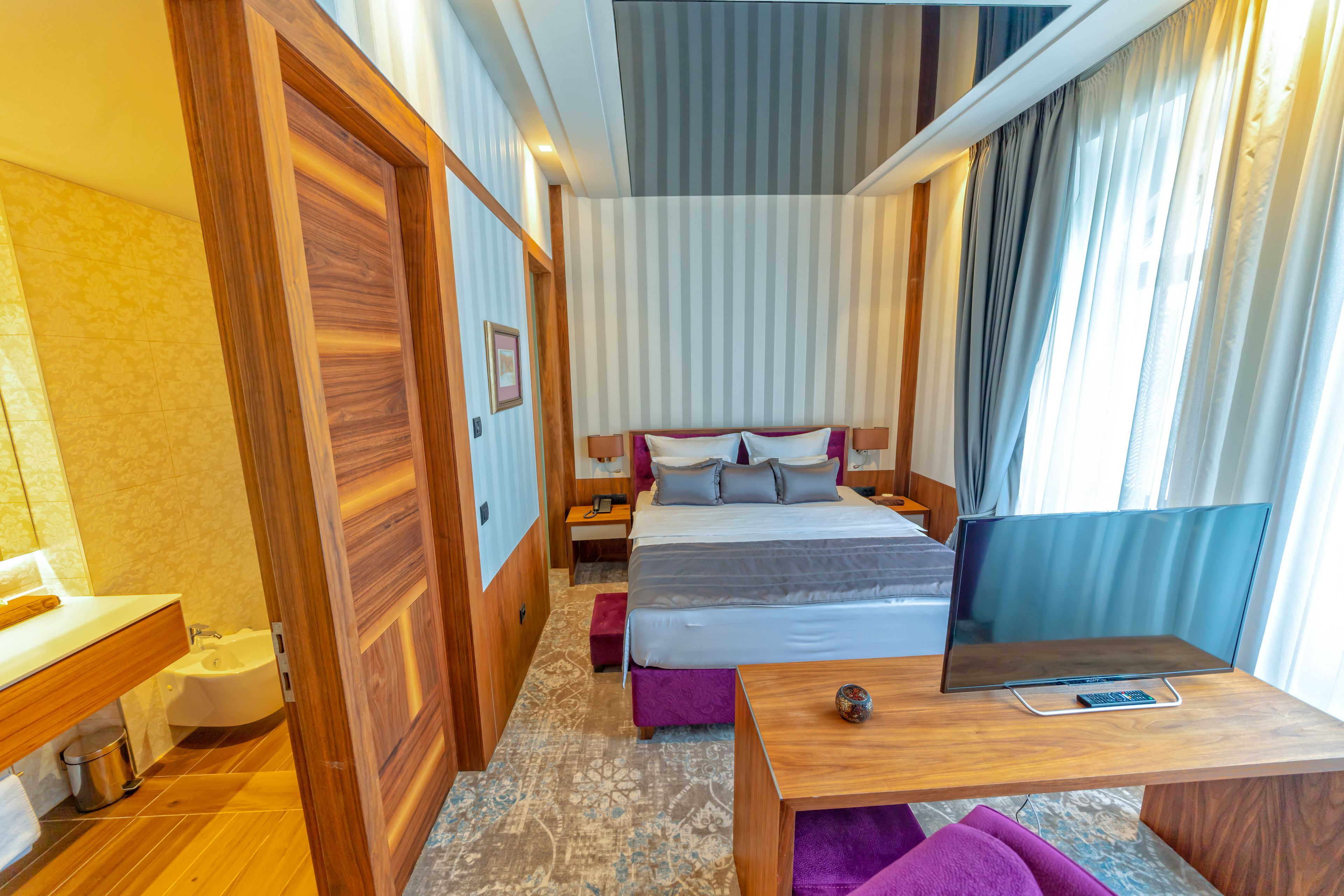 Une chambre d’hôtel avec un plancher en bois, un lit recouvert d’une couette grise et blanche, un bureau en bois, une chaise violette, une table de chevet avec une lampe et une fenêtre avec des rideaux.