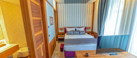 A hotel room with a wooden floor, a bed with a grey and white duvet, a wooden desk, a purple chair, a bedside table with a lamp, and a window with curtains.