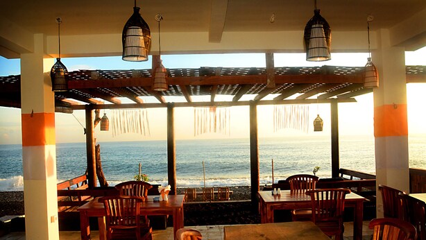 An outdoor seating area with wooden tables and chairs, overlooking the ocean. - Amed Sunset Beach (Karangasem)