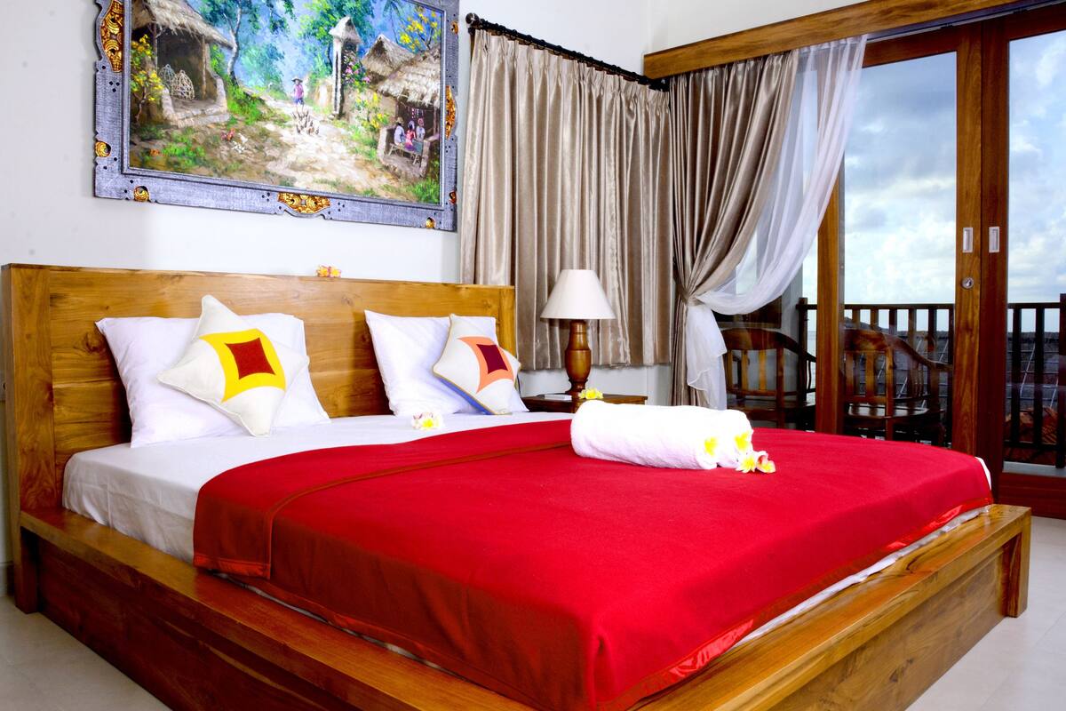 A bed with a red bedspread, wooden headboard, and white bedding. There is a painting on the wall above the bed and a balcony with a railing.