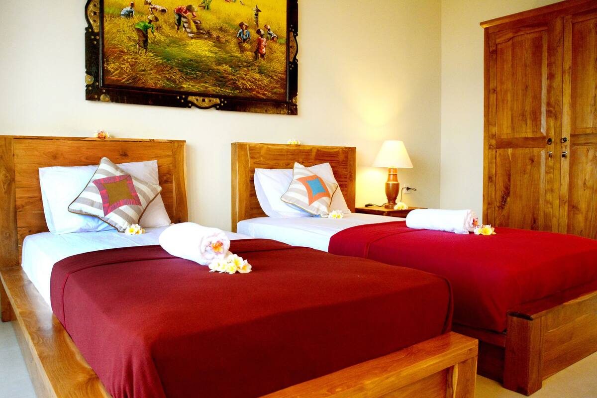 A hotel room with two wooden beds, each with a red bedspread and white linens, a wooden headboard, a nightstand with a lamp, and a large wooden wardrobe.