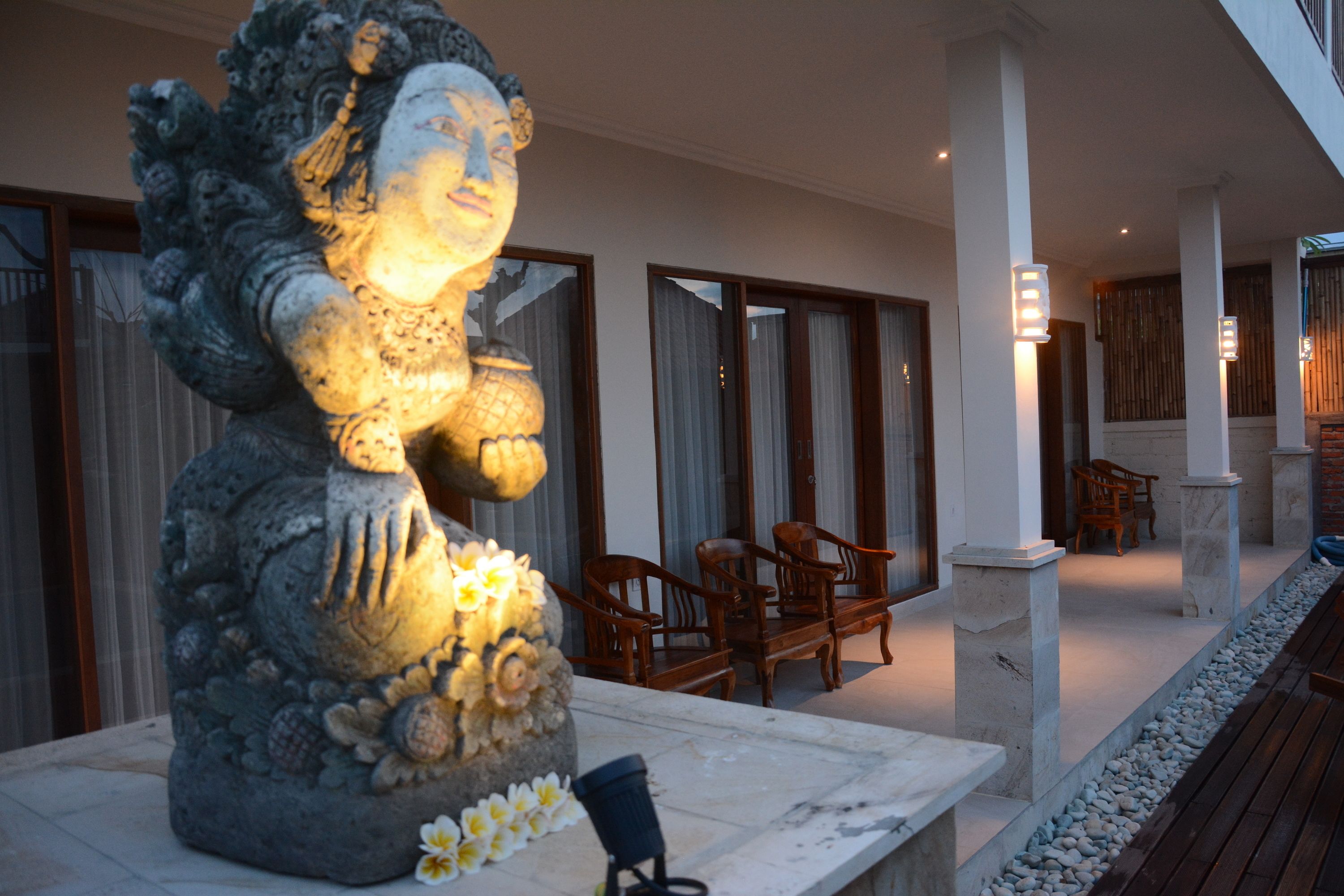 A stone statue with floral decorations on a marble platform, wooden chairs, and a stone pathway in a covered outdoor area.