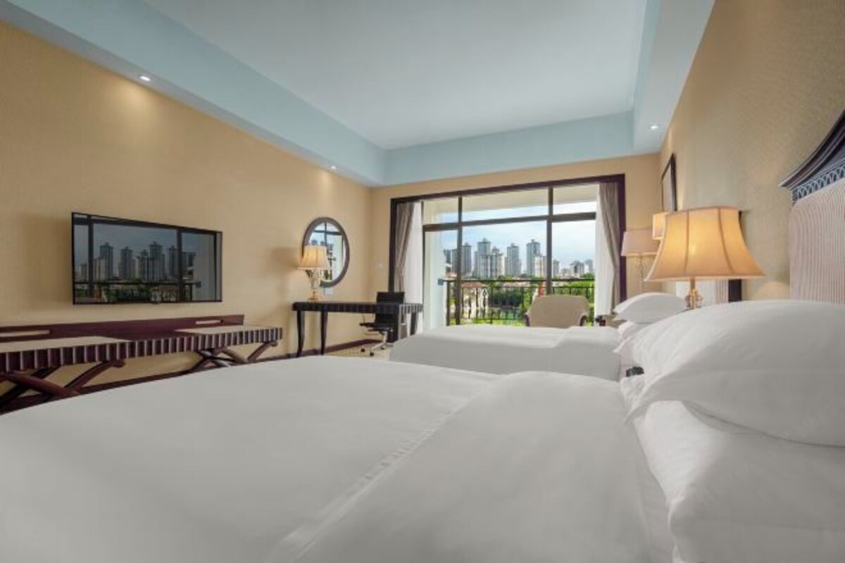 A hotel room with two beds, a desk, a chair, and a large window offering a city view.