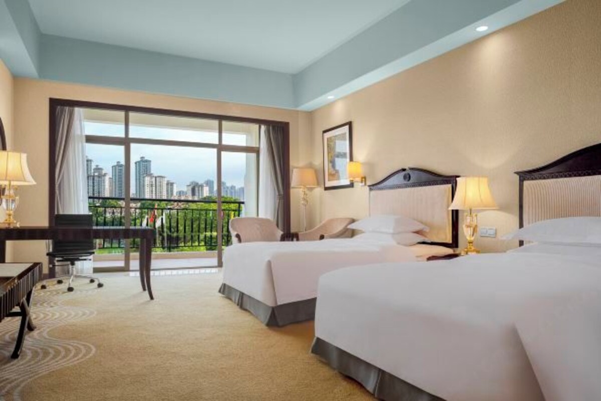 A hotel room with two beds, a desk, and a chair. There is a large window with a view of buildings.