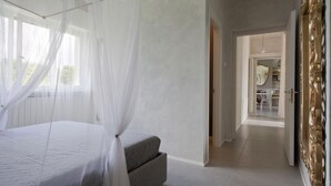 Un dormitori amb un llit, una taula de nit i una finestra amb cortines de tul. Hi ha un passadís que condueix a una zona de dinar.