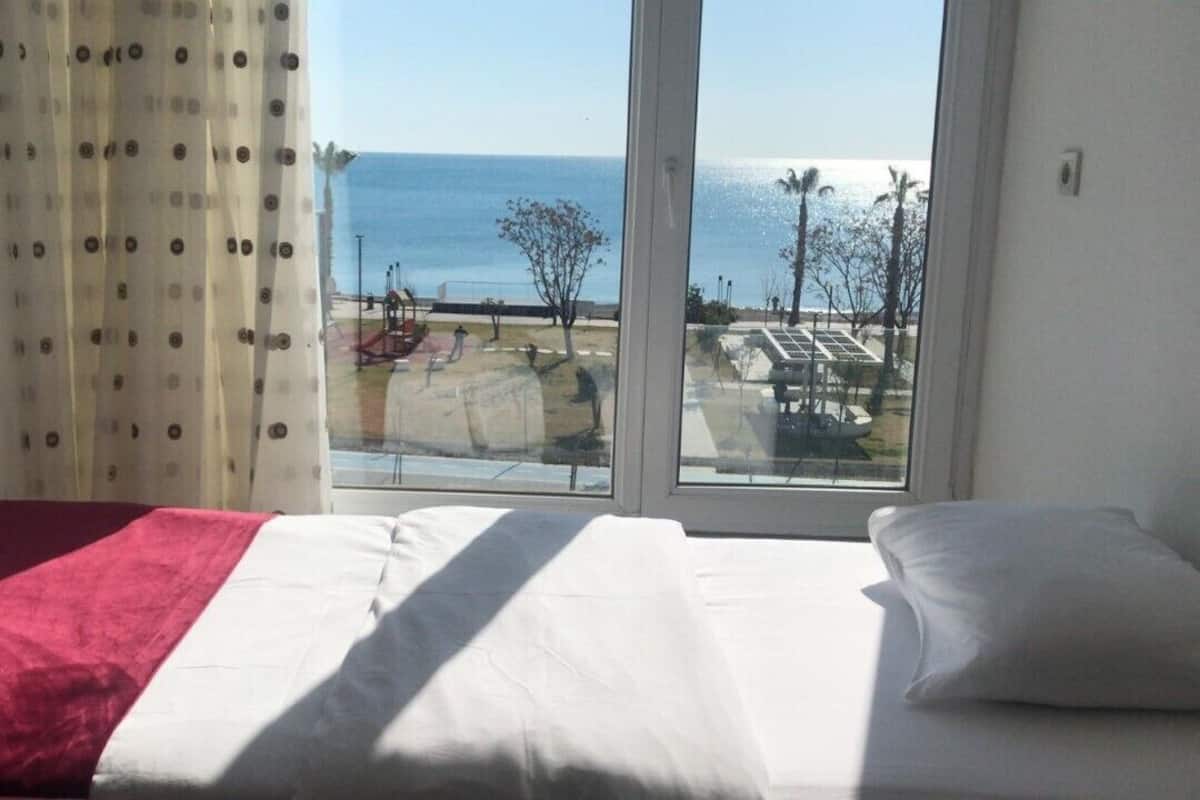 A bed with a red and white bedspread, a white pillow, and a view of a beach and solar panels through a window.