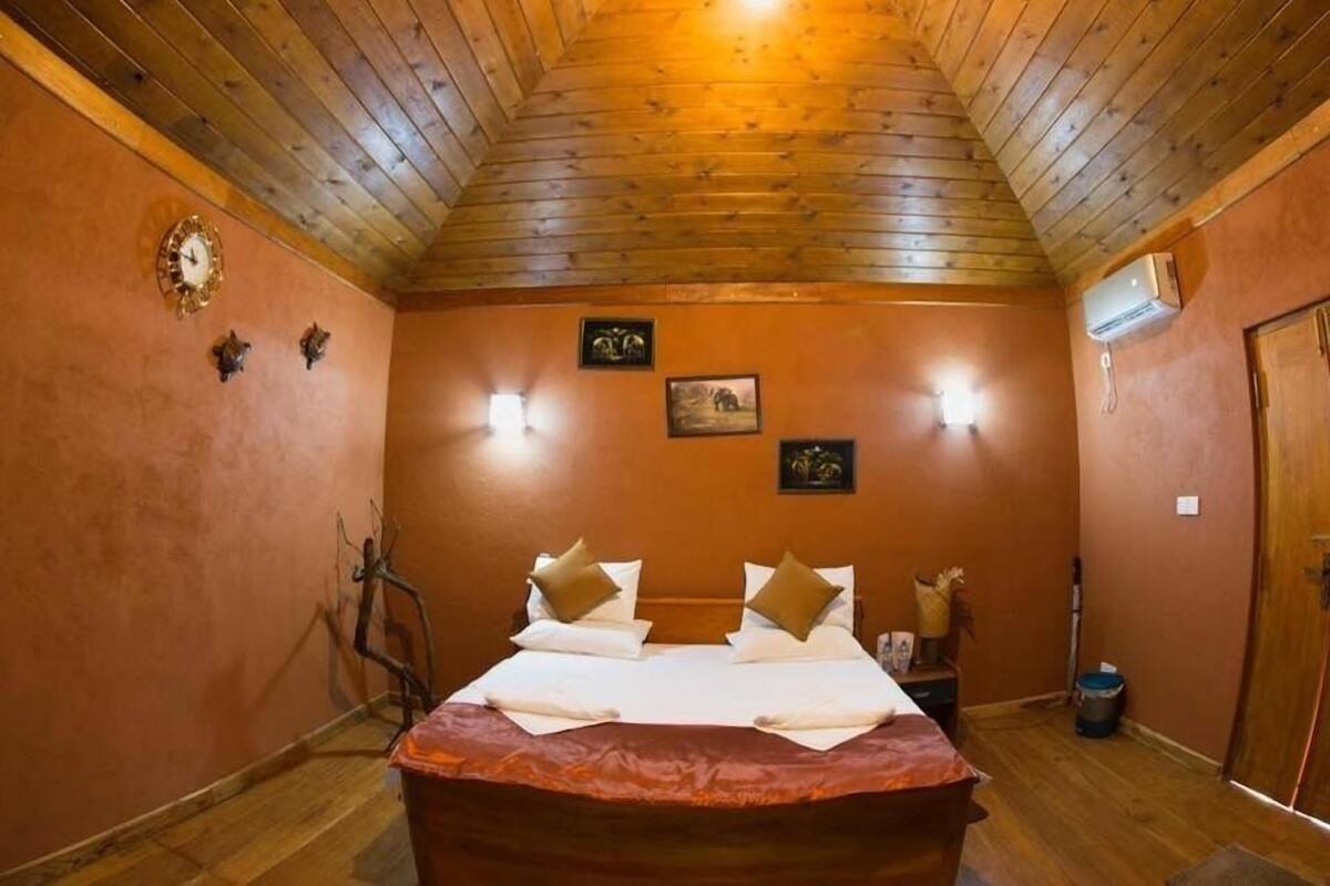 A room with a wooden ceiling, a bed with white linens and two pillows, a wall clock, and framed pictures.