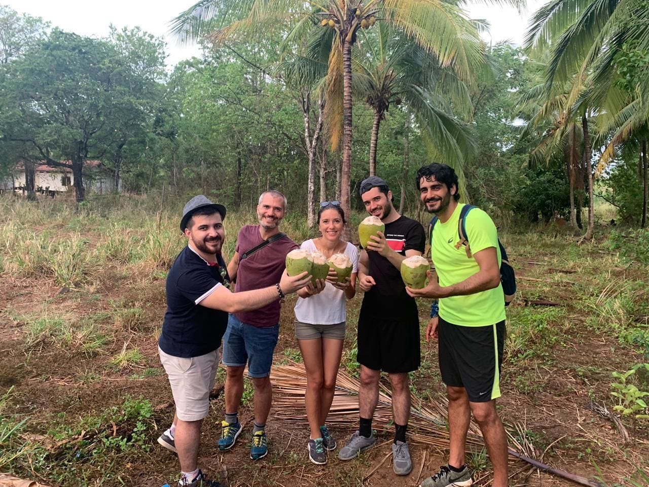A group of people holding coconuts in a natural setting with trees and a dirt path.