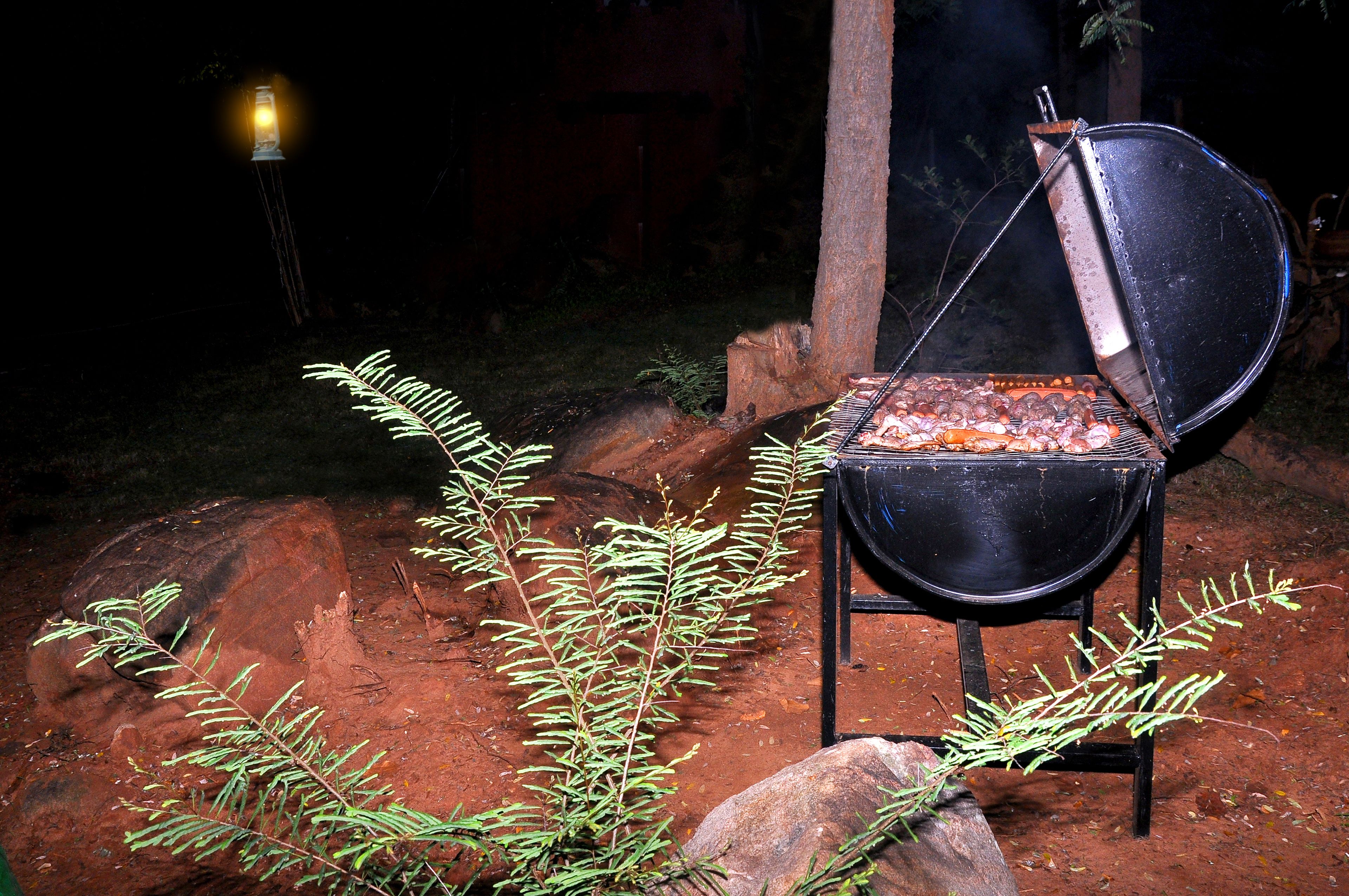 An outdoor grill with a black metal frame and a lid, set on a dirt ground with rocks and green foliage around.