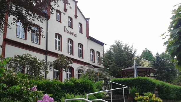 Een gebouw met de naam 'Vesta' op de gevel, omringd door weelderige begroeiing en bloeiende planten.