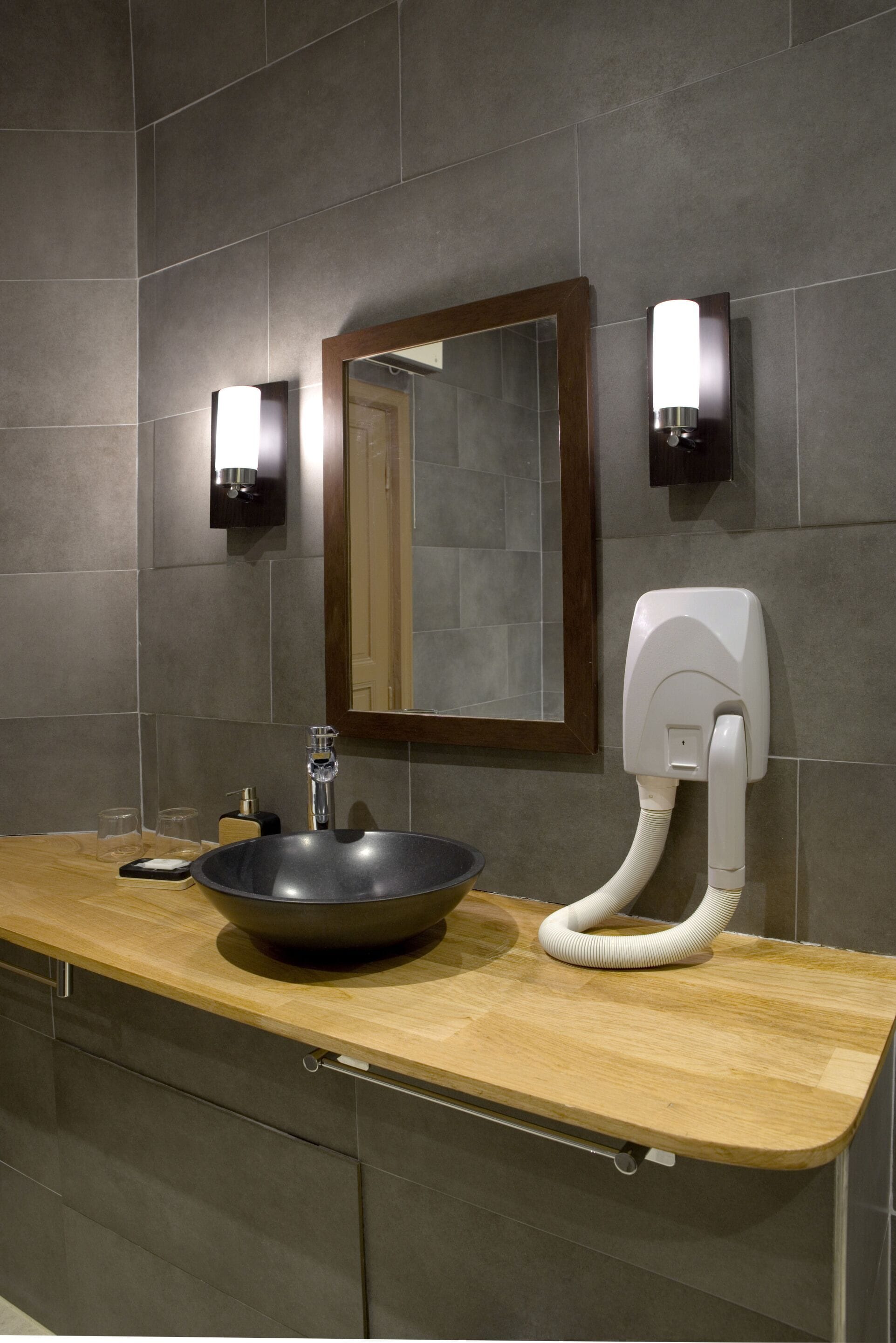A modern bathroom with a wooden vanity, a black vessel sink, a mirror with a wooden frame, and two wall-mounted light fixtures.