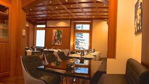 A dining area with a glass table, chairs, and a bench. There are windows with artwork and a wooden ceiling. - arthausHOTEL (Davos)